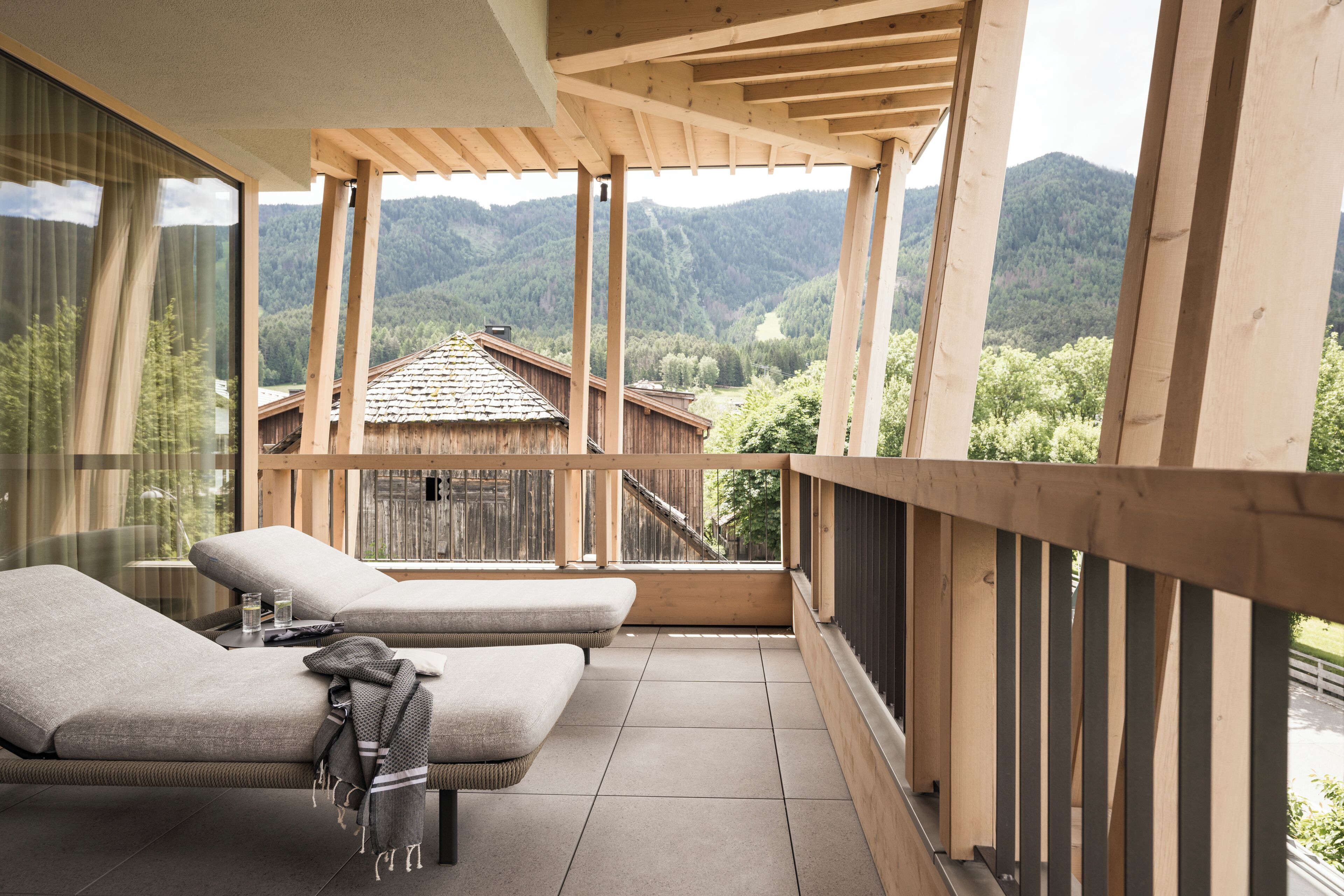 A balcony with two cushioned seating areas, a view of a mountain landscape, and a wooden pergola structure.