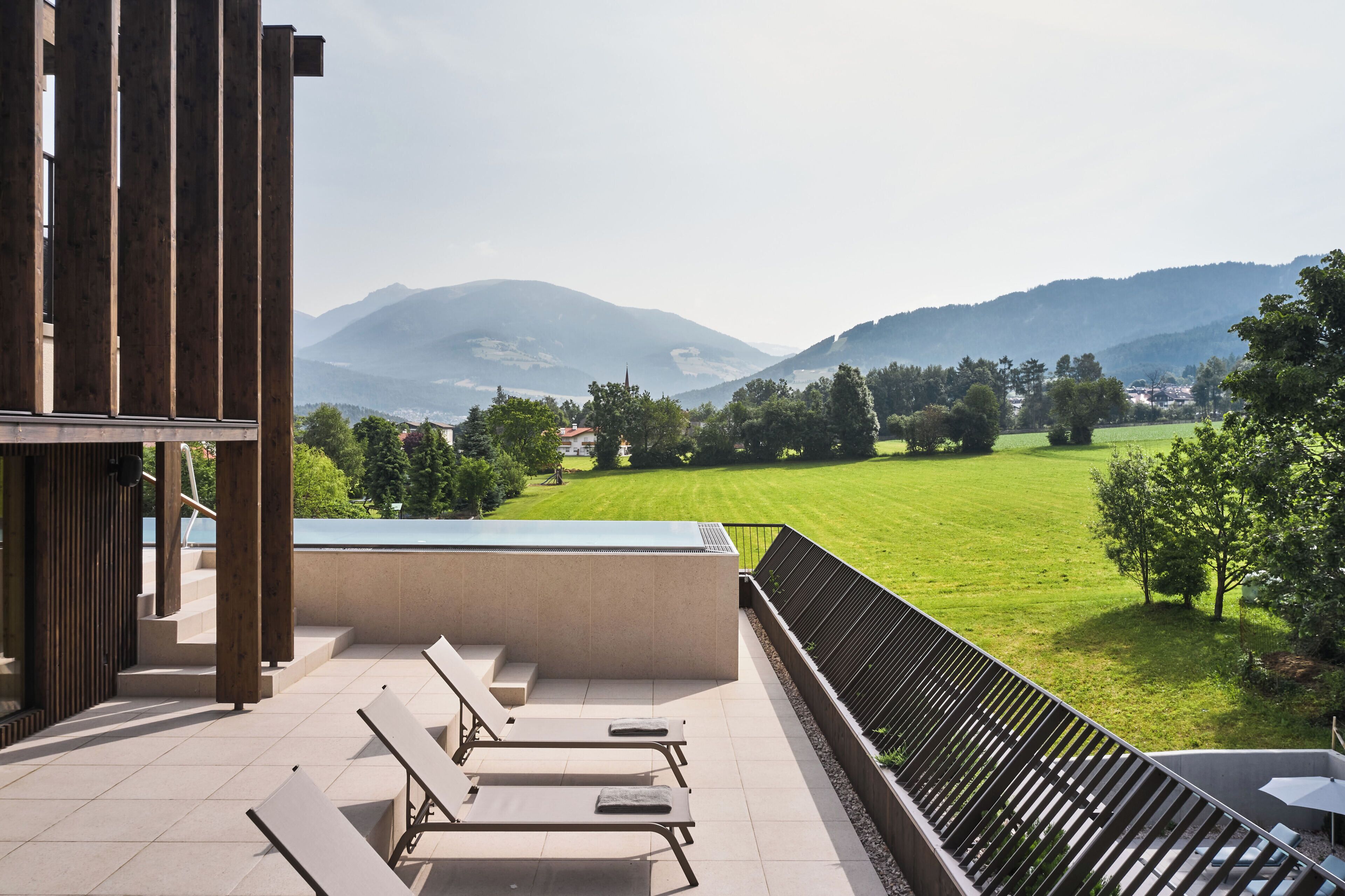 A terrace with lounge chairs, a view of a green field, and mountains in the distance.