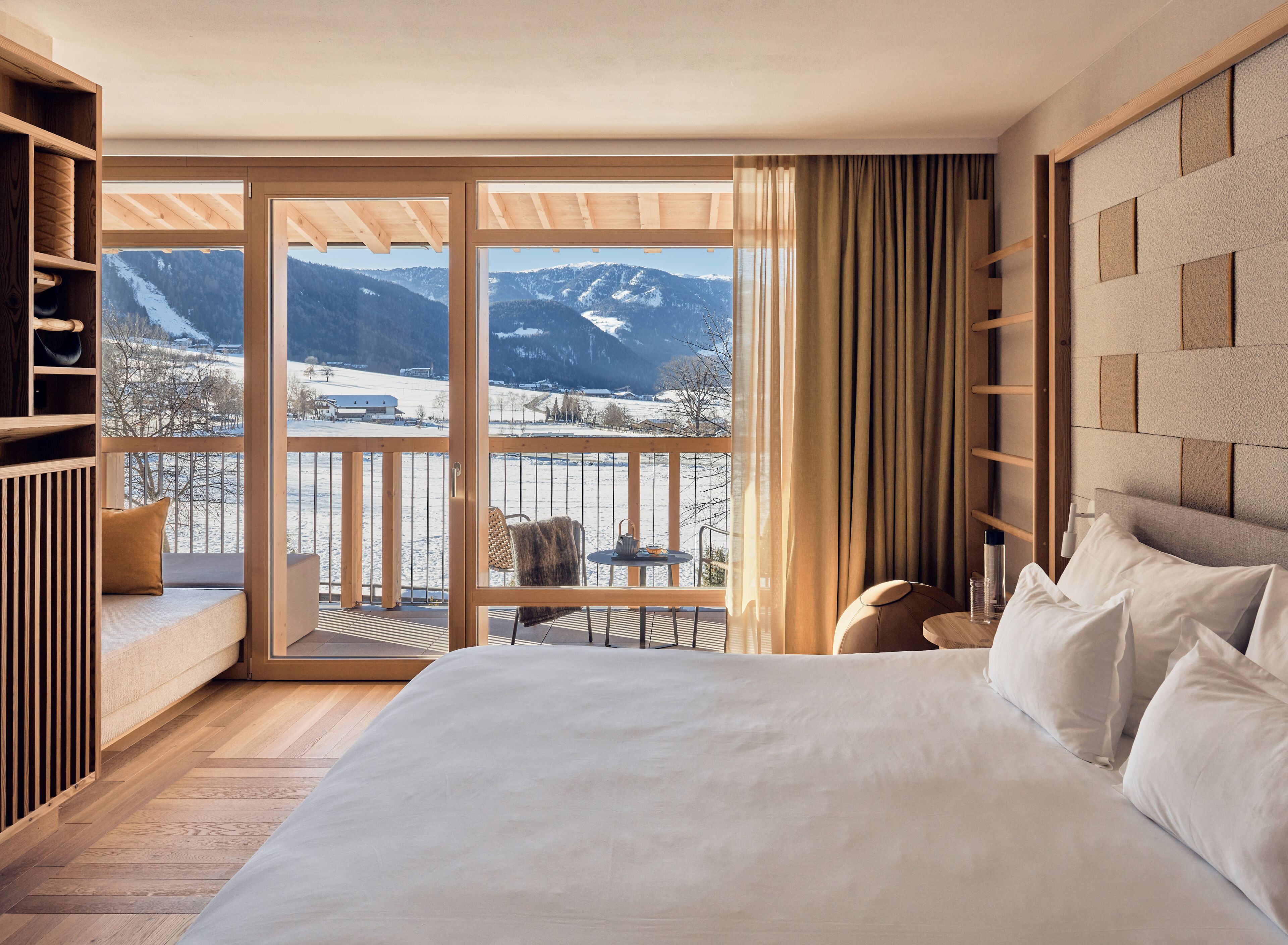 A bedroom with a large bed, a wooden headboard, a balcony with a view of snow-covered mountains, and a seating area with a table and chairs.