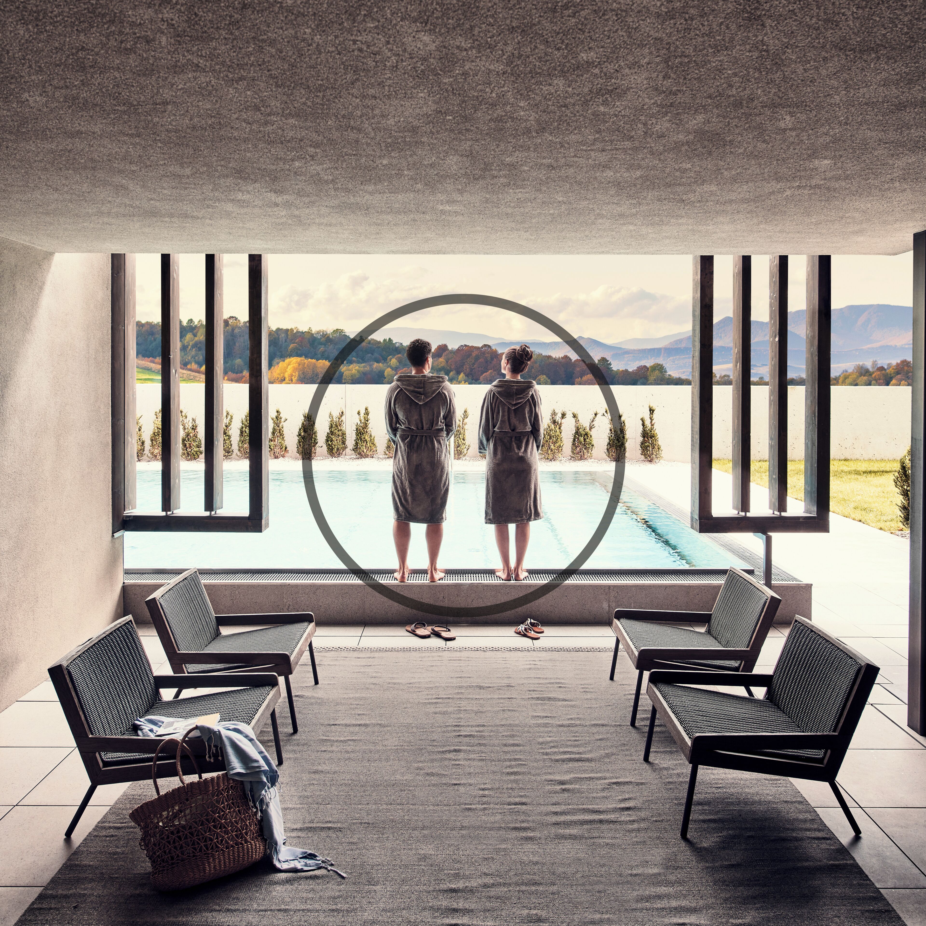A room with a large window overlooking a lake, two people standing by the pool, and seating area with chairs and a basket.