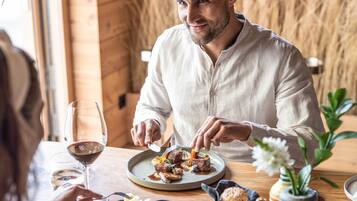 Um homem jantando em uma mesa de madeira, com um prato de comida, um copo de vinho e um vaso com flores.