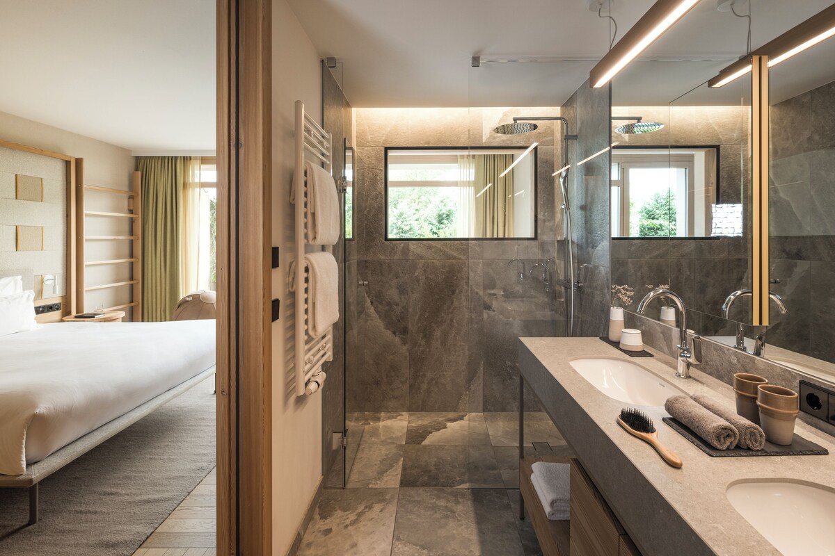 Una habitación de hotel moderna con una cama grande, un baño con ducha de vidrio y un tocador con espejo.