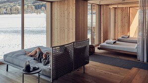 A modern room with a large window overlooking a snowy landscape, a sofa, a bed, and a small table with a bag. - Falkensteiner Hotel Kronplatz - The Leading Hotels of the World (Brunico)