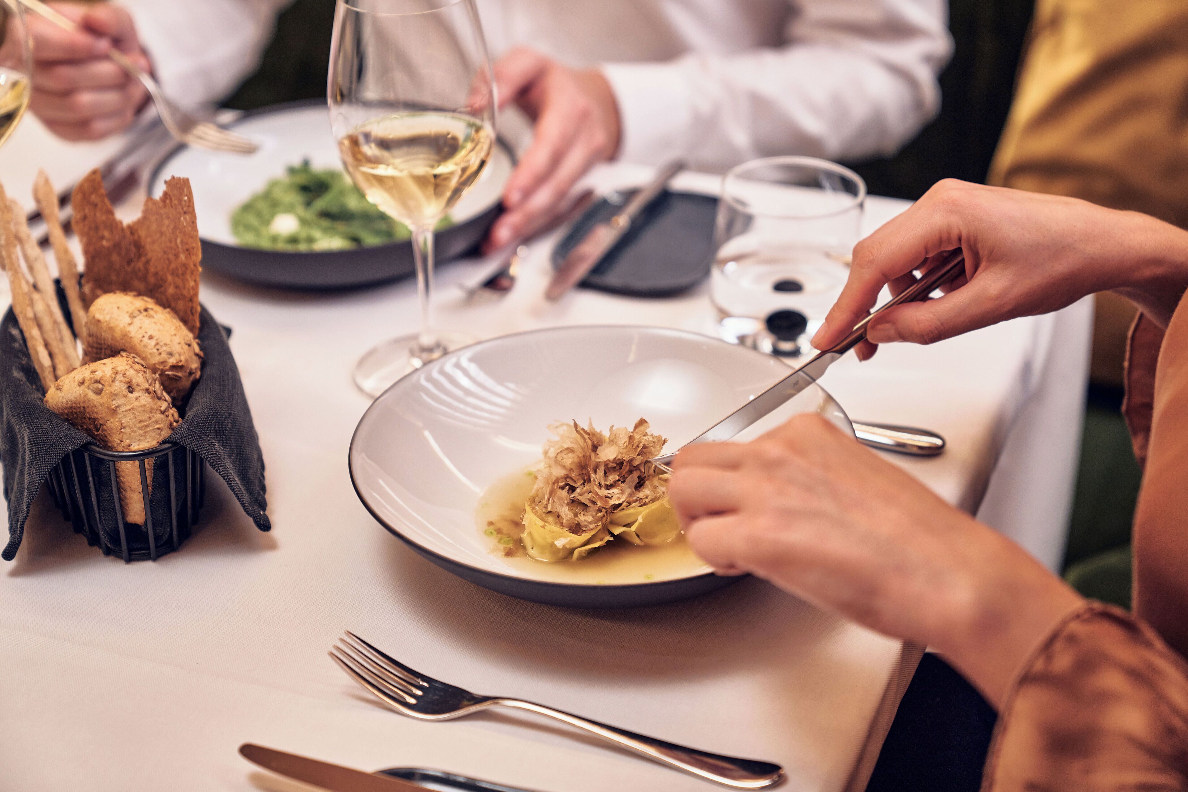 Una mesa de comedor con un plato de comida, cubiertos y una copa de vino blanco.
