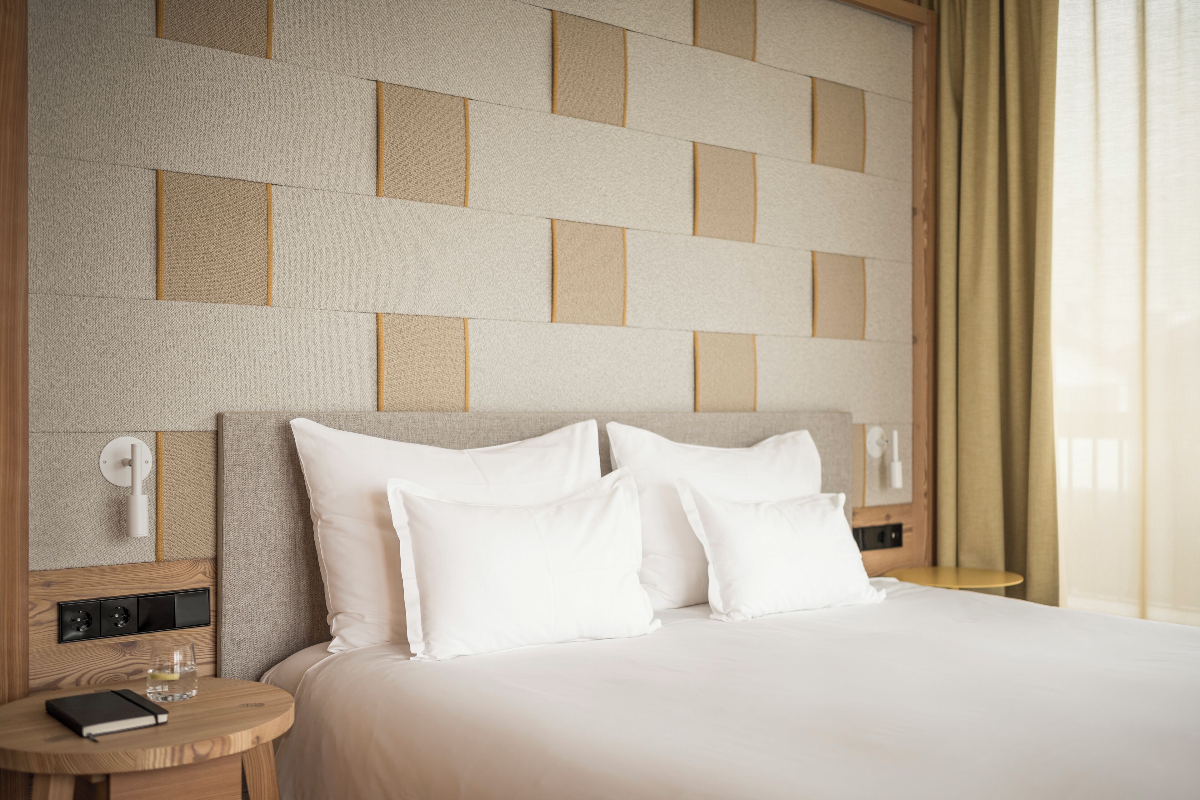 A neatly made bed with white linens, a bedside table with a book and glasses, and a wall with a geometric pattern.