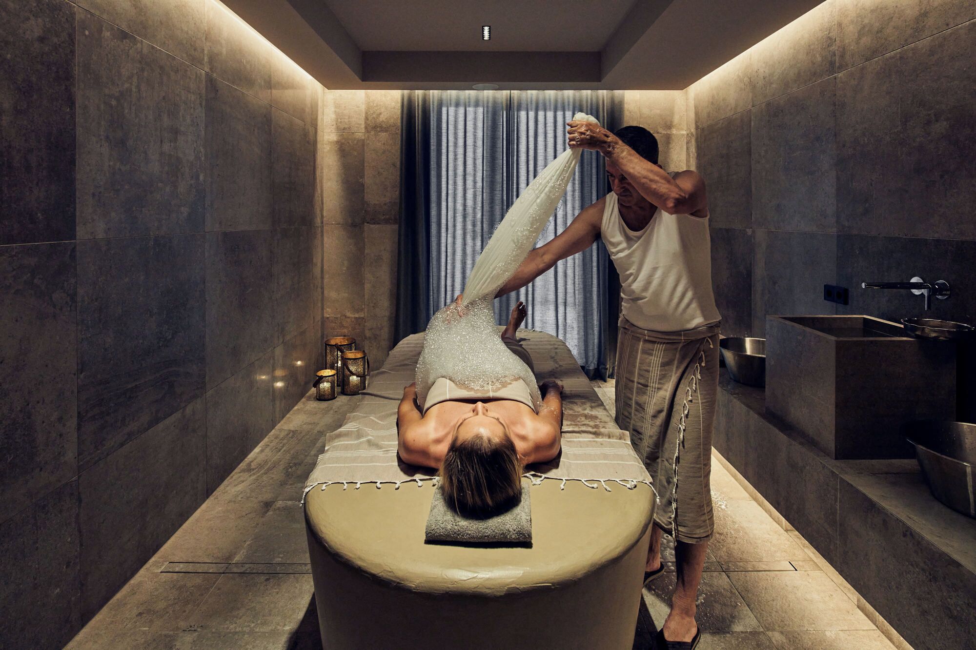 Un cuarto de spa con una camilla de masaje, una persona recibiendo un masaje en la espalda y un lavamanos pequeño.
