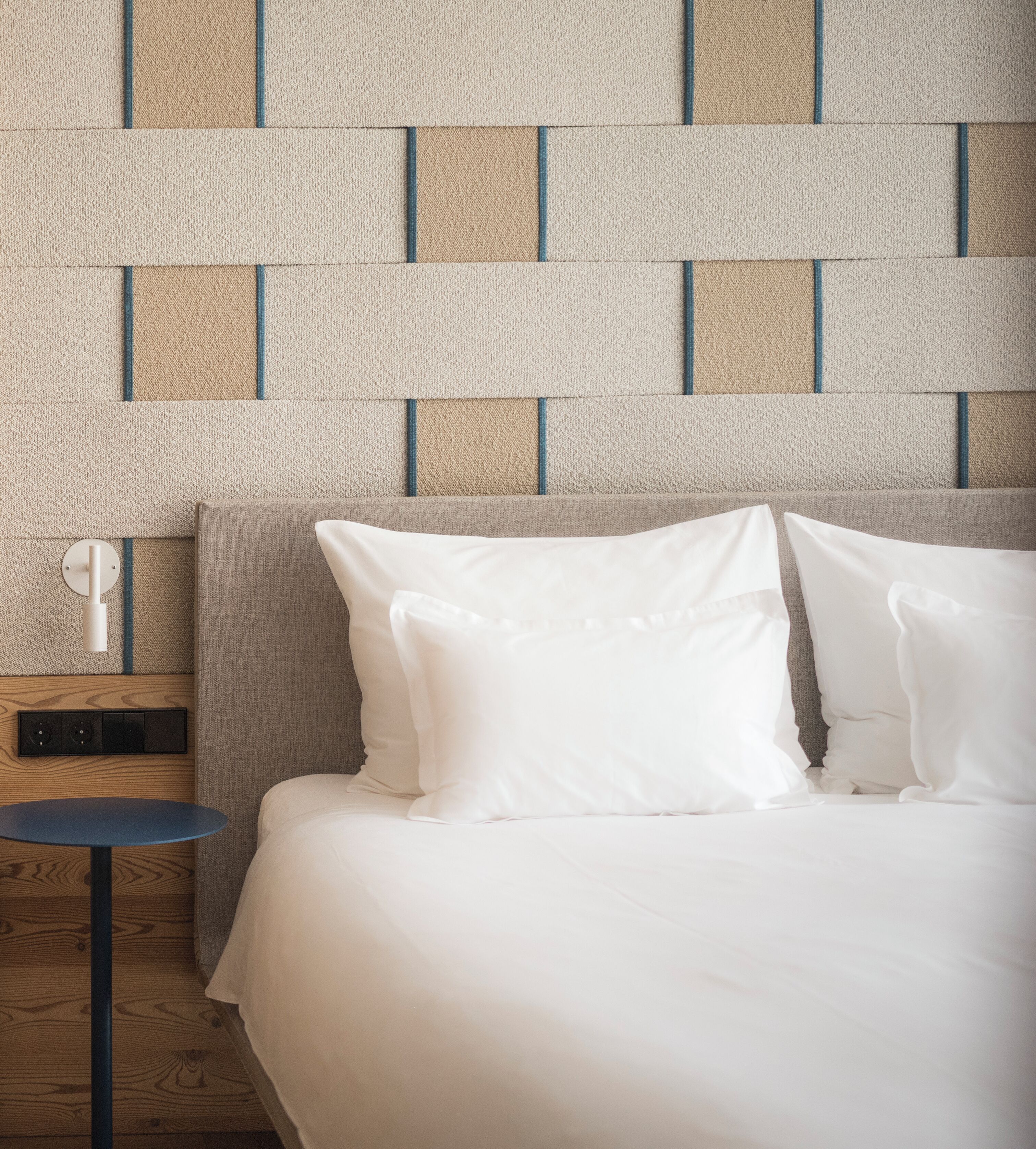 A neatly made bed with white linens, a wooden nightstand with a lamp, and a wall with a geometric pattern.