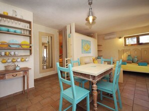 Uma sala de jantar com mesa de madeira e cadeiras cor turquesa, uma cozinha com prateleiras cheias de louças e uma cama com cabeceira amarela.