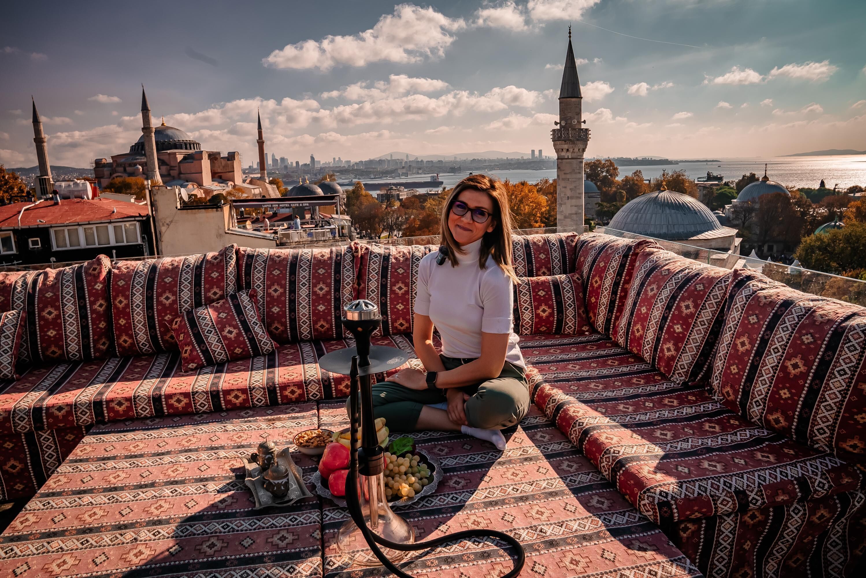 Iemand zit op een gestreept bankje met een shisha en fruit op een tafel, met uitzicht op een stadsbeeld met koepels en een rivier.