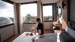 A modern hotel room with a large window offering a view of a cityscape, including a mosque. There is a bed with a patterned bedspread, a desk with a chair, and a small table with a plate of pastries. - World Heritage Center Hotel (Istanbul)