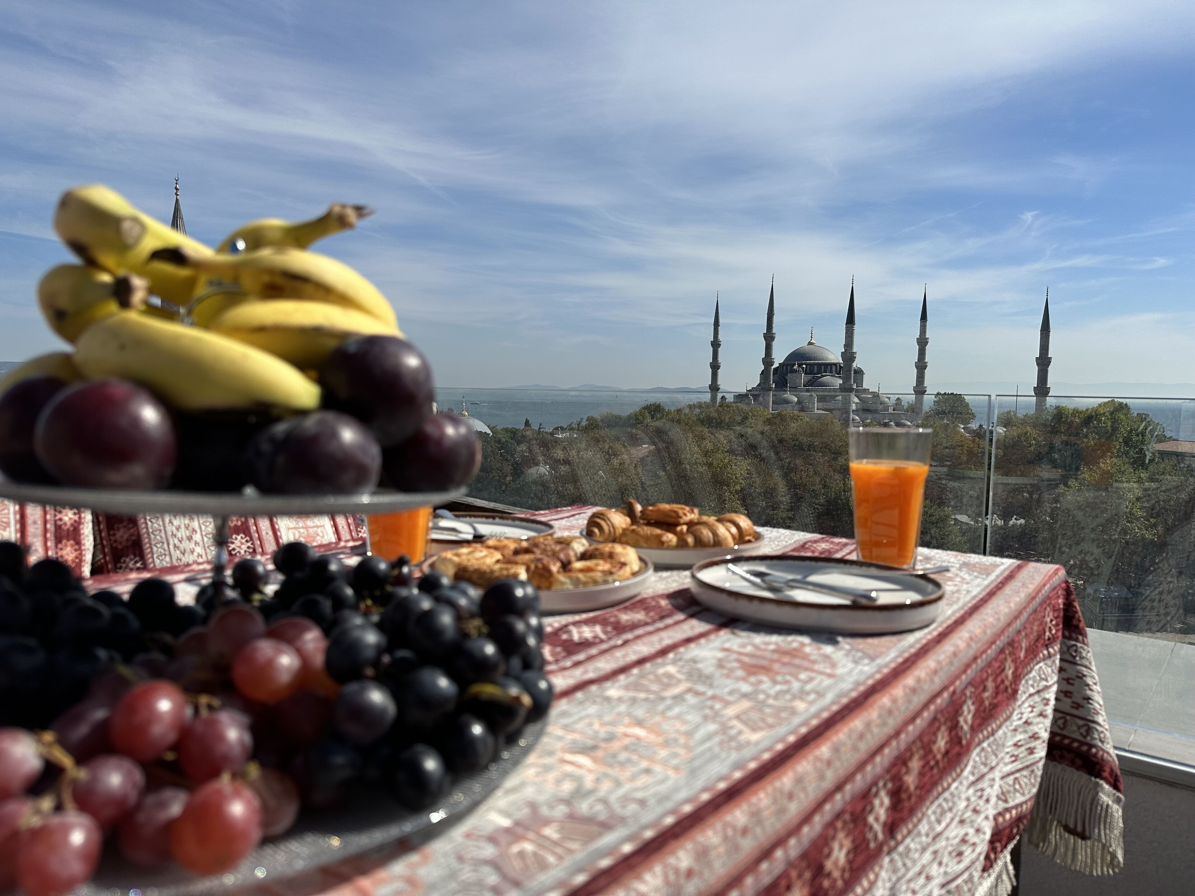 A table with fruits, pastries, and a glass of orange juice, overlooking a view of a mosque with four minarets.