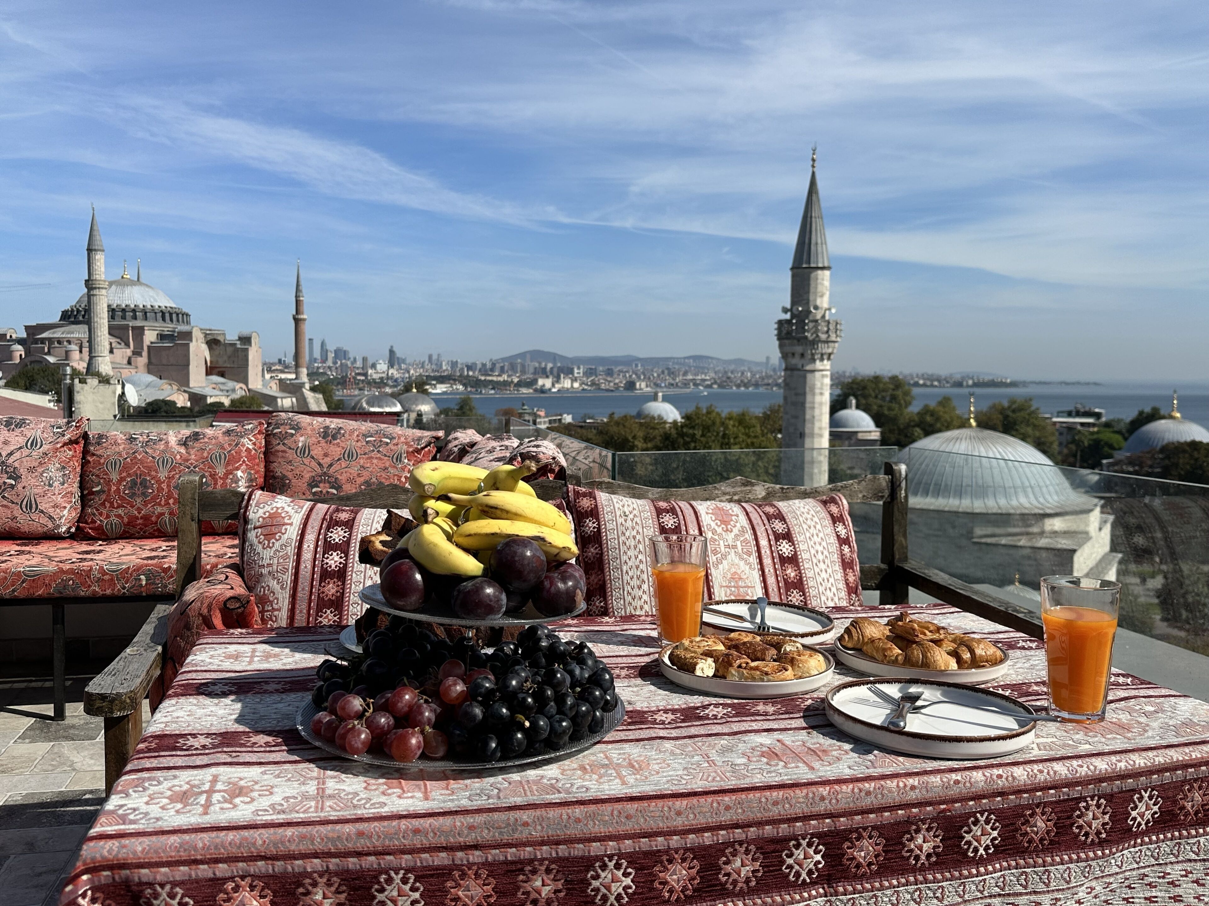 A table with a fruit platter, croissants, and juice glasses, overlooking a cityscape with domed buildings and a minaret.