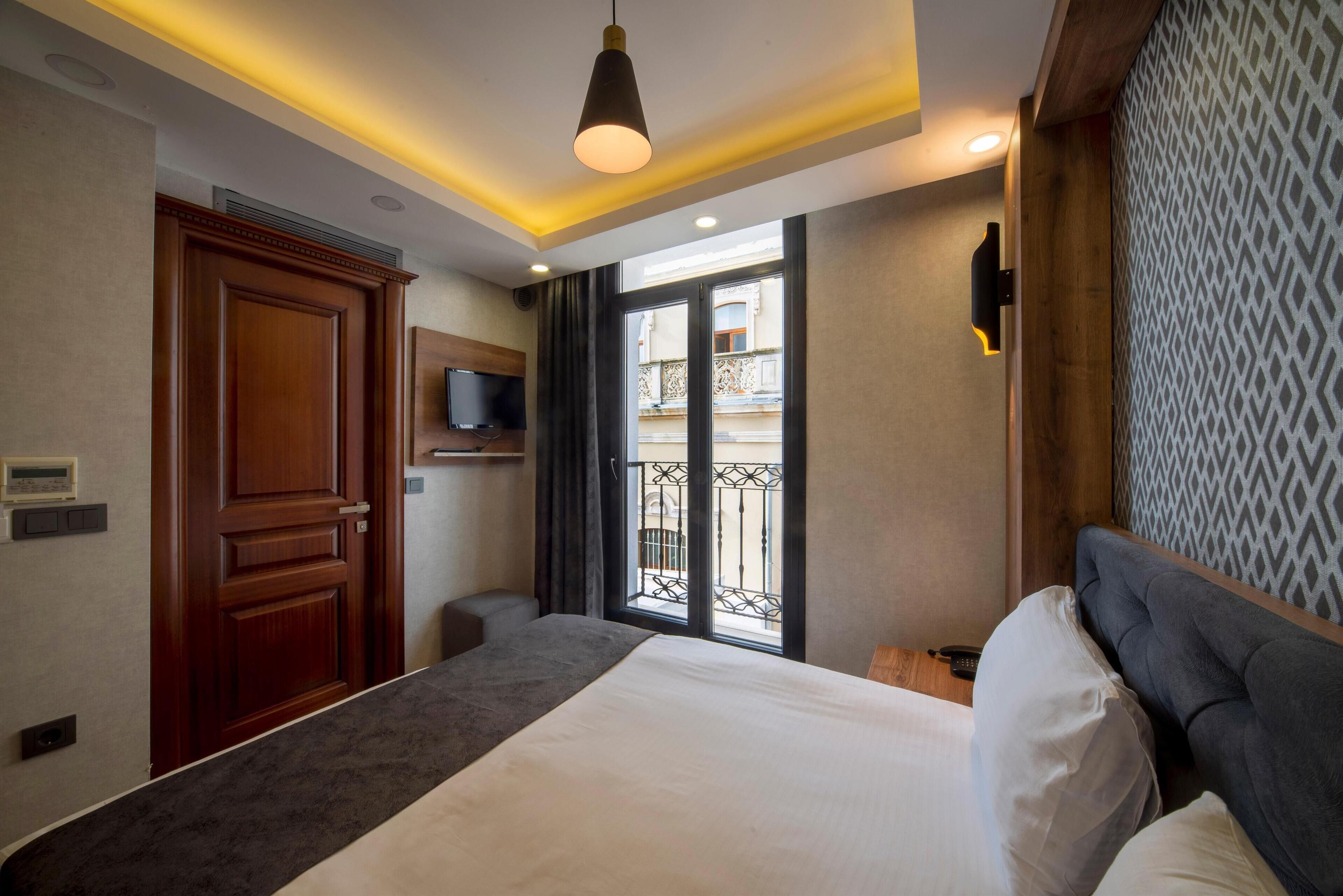 A hotel room with a large bed, a TV mounted on the wall, a wooden wardrobe, and a balcony with a wrought iron gate.