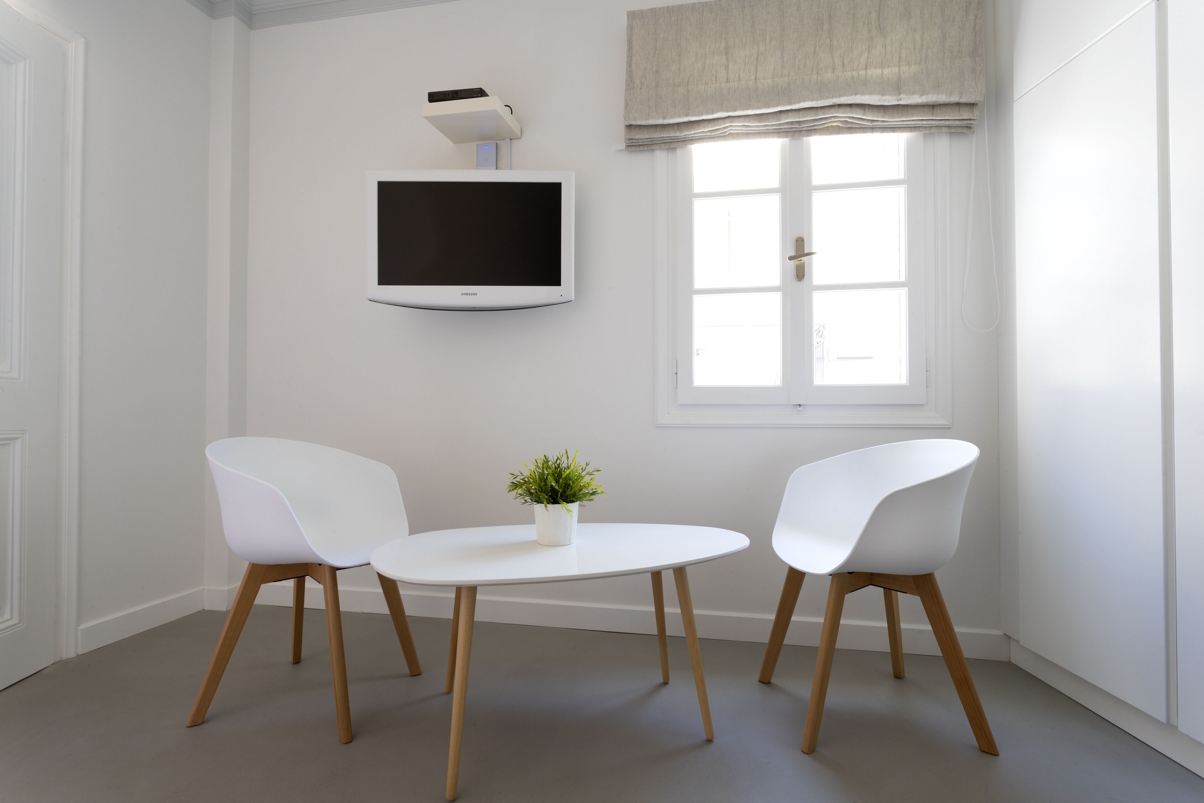A room with a white wall-mounted TV, a window with a valance, and two white chairs around a small table with a potted plant.
