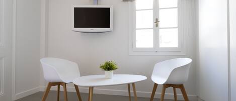 A room with a white wall-mounted TV, a window with a valance, and two white chairs around a small table with a potted plant.