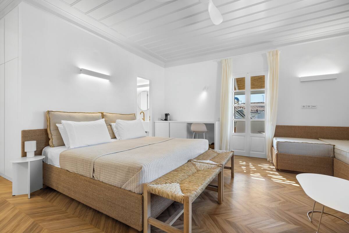 A modern bedroom with a large bed, a bench, a desk, and a view of the outside.