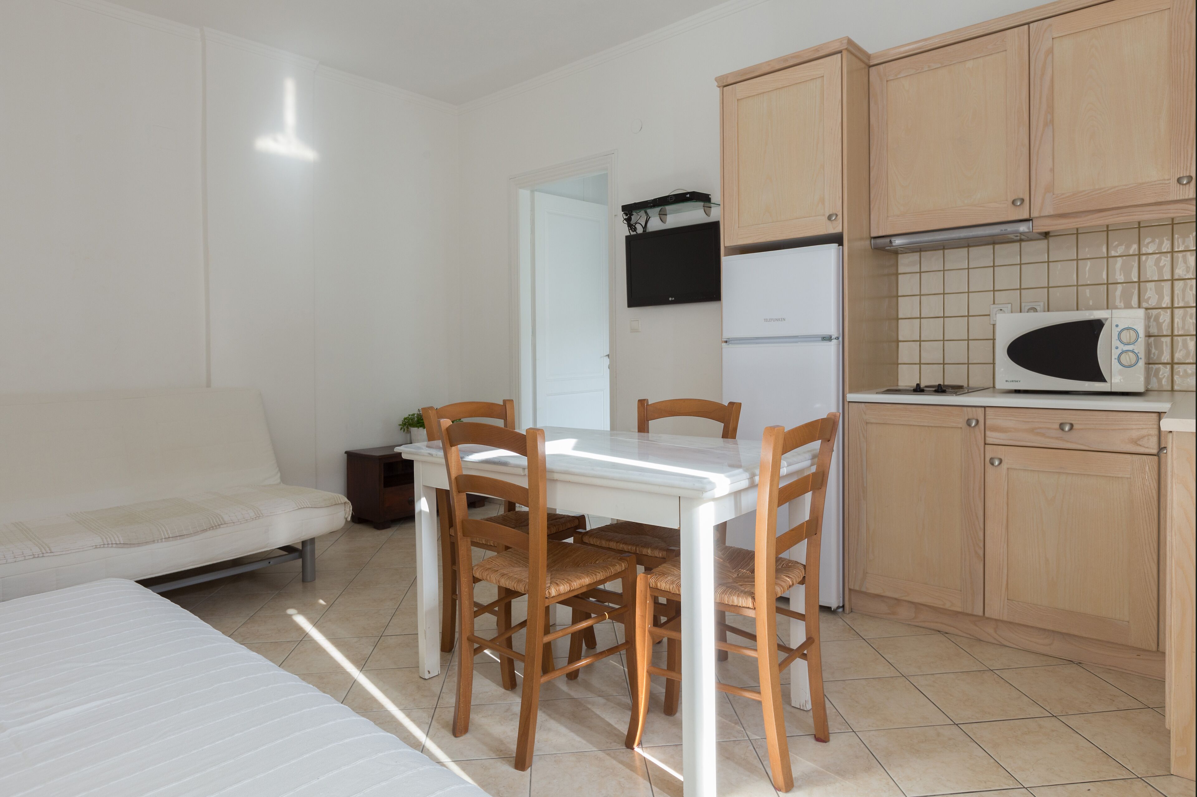 A small, clean room with a bed, a dining table with chairs, a kitchenette with a microwave, and a refrigerator.