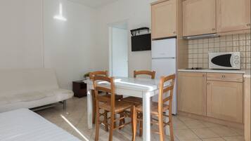 A small, clean room with a bed, a dining table with chairs, a kitchenette with a microwave, and a refrigerator.