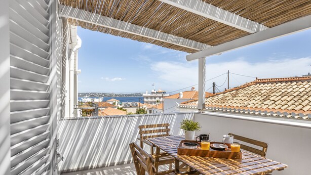 Un balcon avec une table dressée pour deux personnes, une plante en pot et une vue sur la mer et les bâtiments.