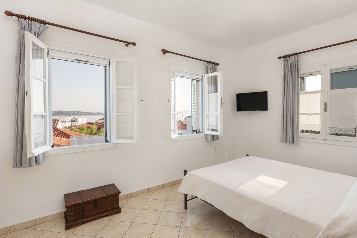 A bedroom with a bed, two windows with curtains, a wooden bench, and a television on the wall.