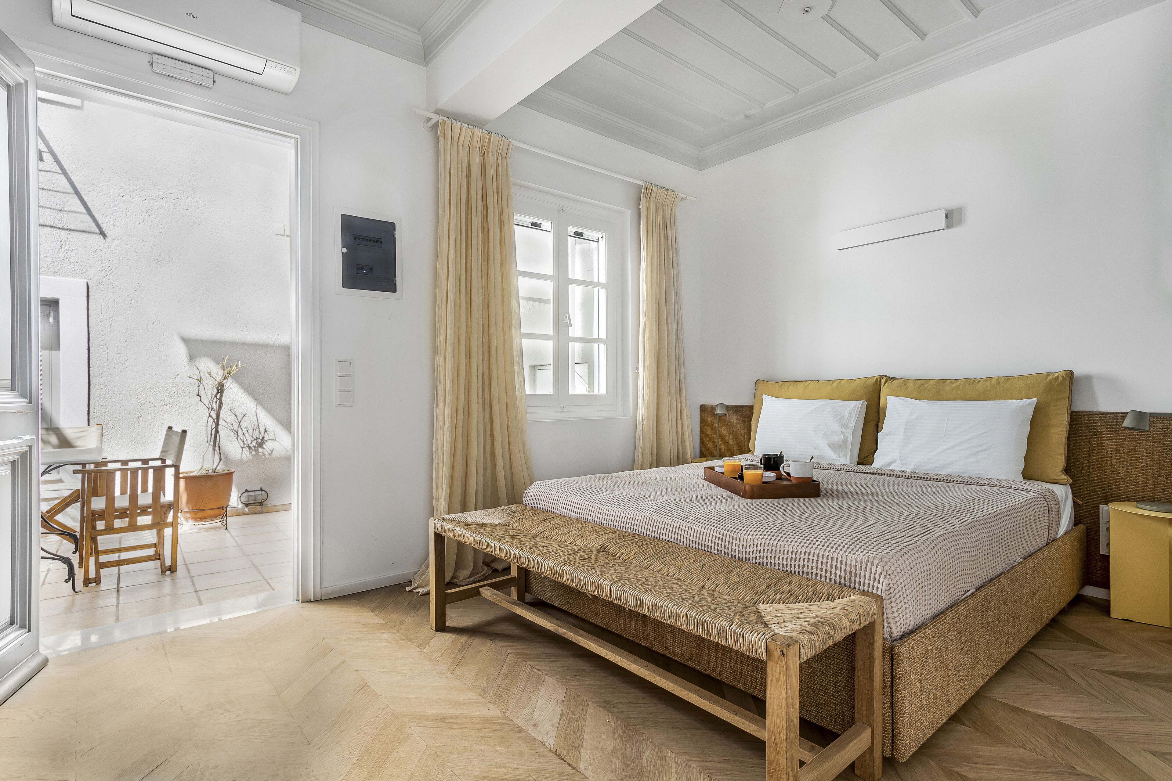 Un dormitorio con cama, mesita de noche, banco y vistas a un balcón.