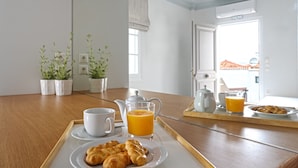 Un plateau de petit-déjeuner avec des croissants, du jus d’orange et une théière, posé sur une table en bois dans une pièce lumineuse aux murs blancs et agrémentée de plantes en pot.