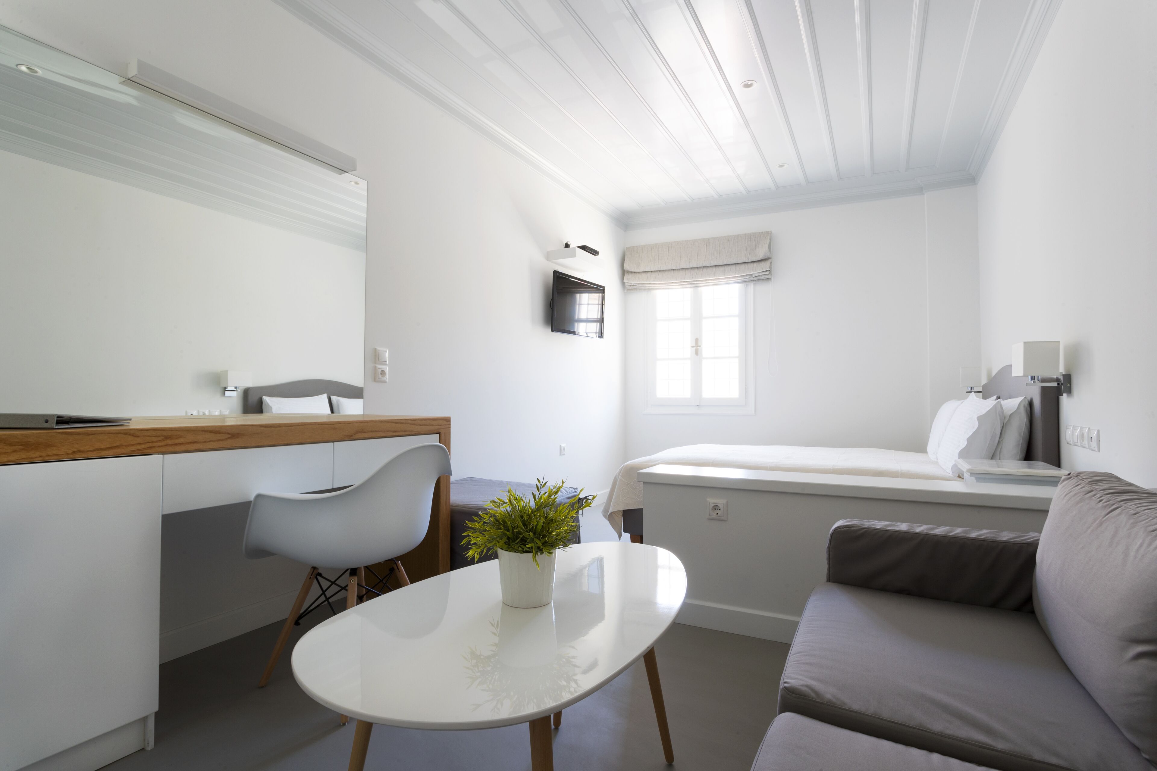 A modern, well-lit room with a dining area, a small table with a plant, a grey sofa, and a bed with a window view.