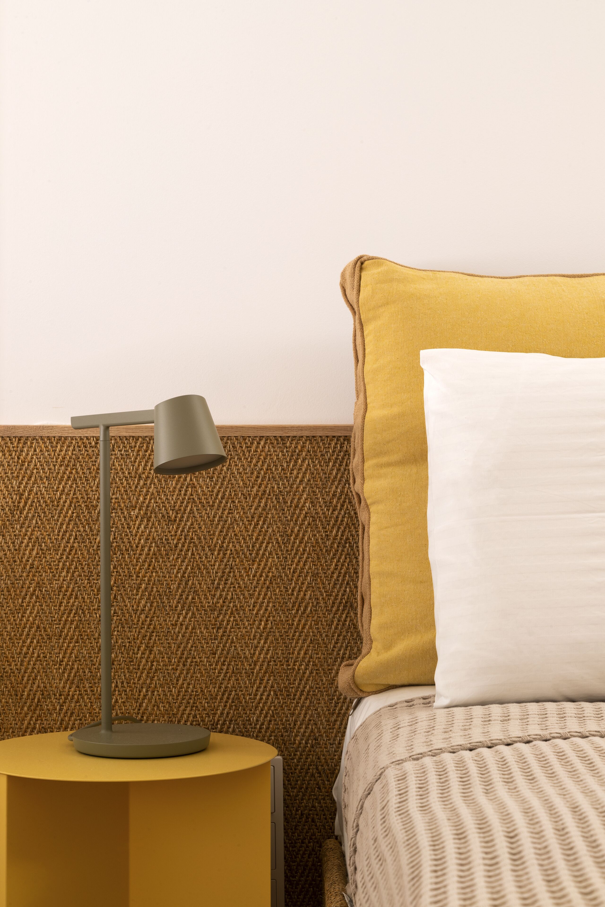 A bed with a mustard-colored pillow and a white pillow, a round yellow nightstand with a lamp, and a herringbone-patterned wall.