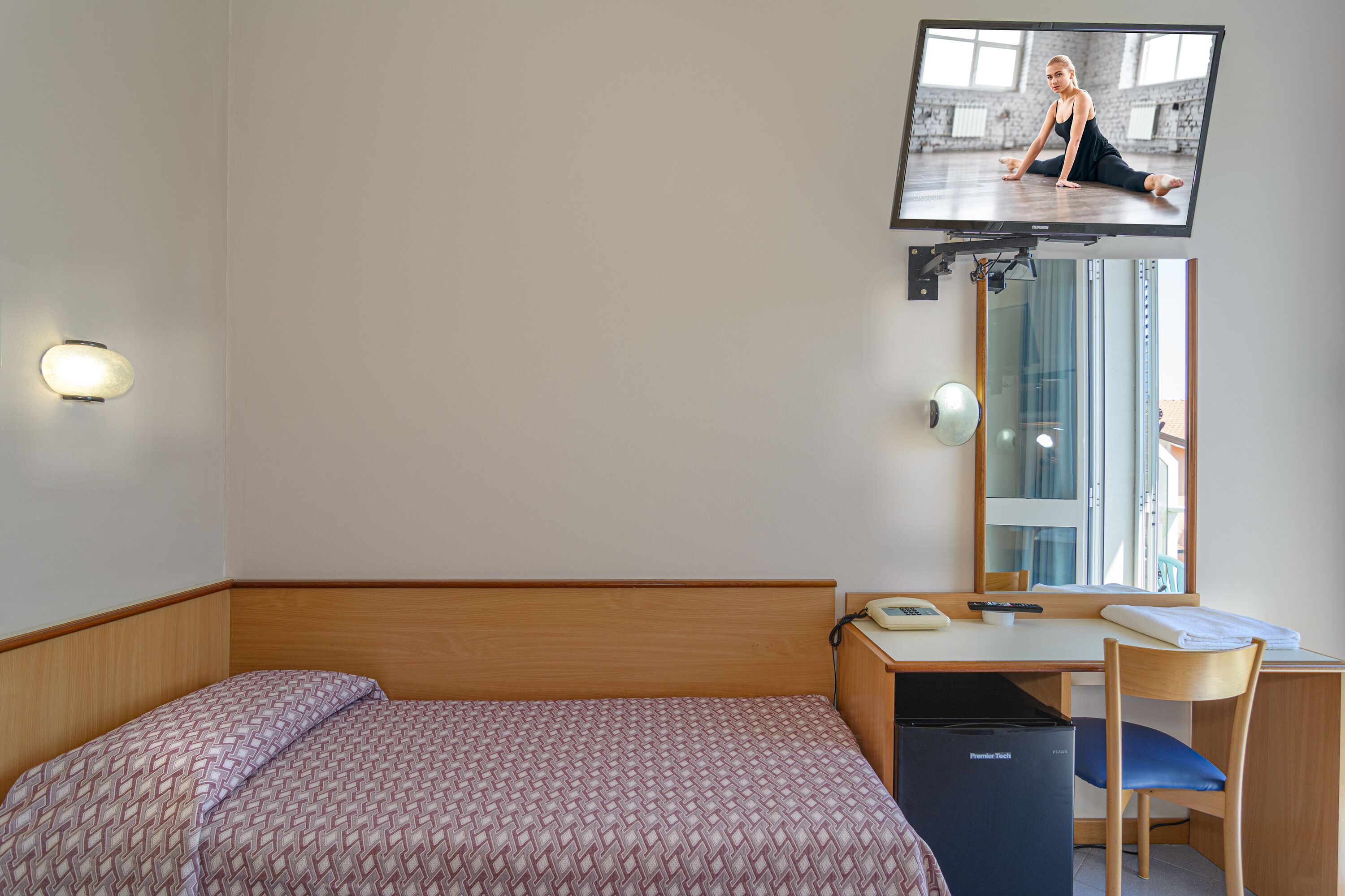 Hotellihuone, jossa on sänky, työpöytä, tuoli, seinälle asennettu televisio ja pieni jääkaappi.