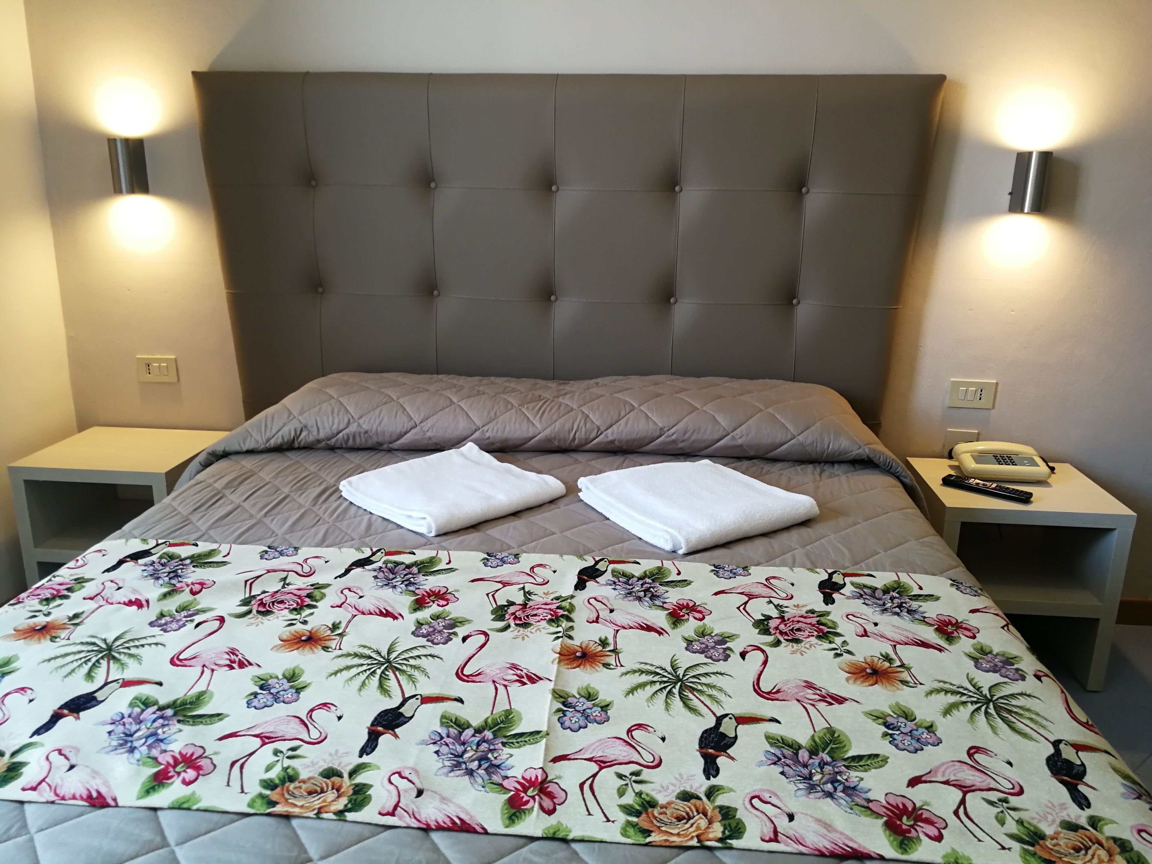 Cama arrumada com colcha estampada em floral, duas toalhas brancas e cabeceira estofada. Há duas mesas de cabeceira, uma com abajur e outra com telefone.