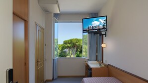 A hotel room with a bed, a desk with a chair, a television mounted on the wall, and a window with a view of trees. - Hotel Sogno (Viareggio)
