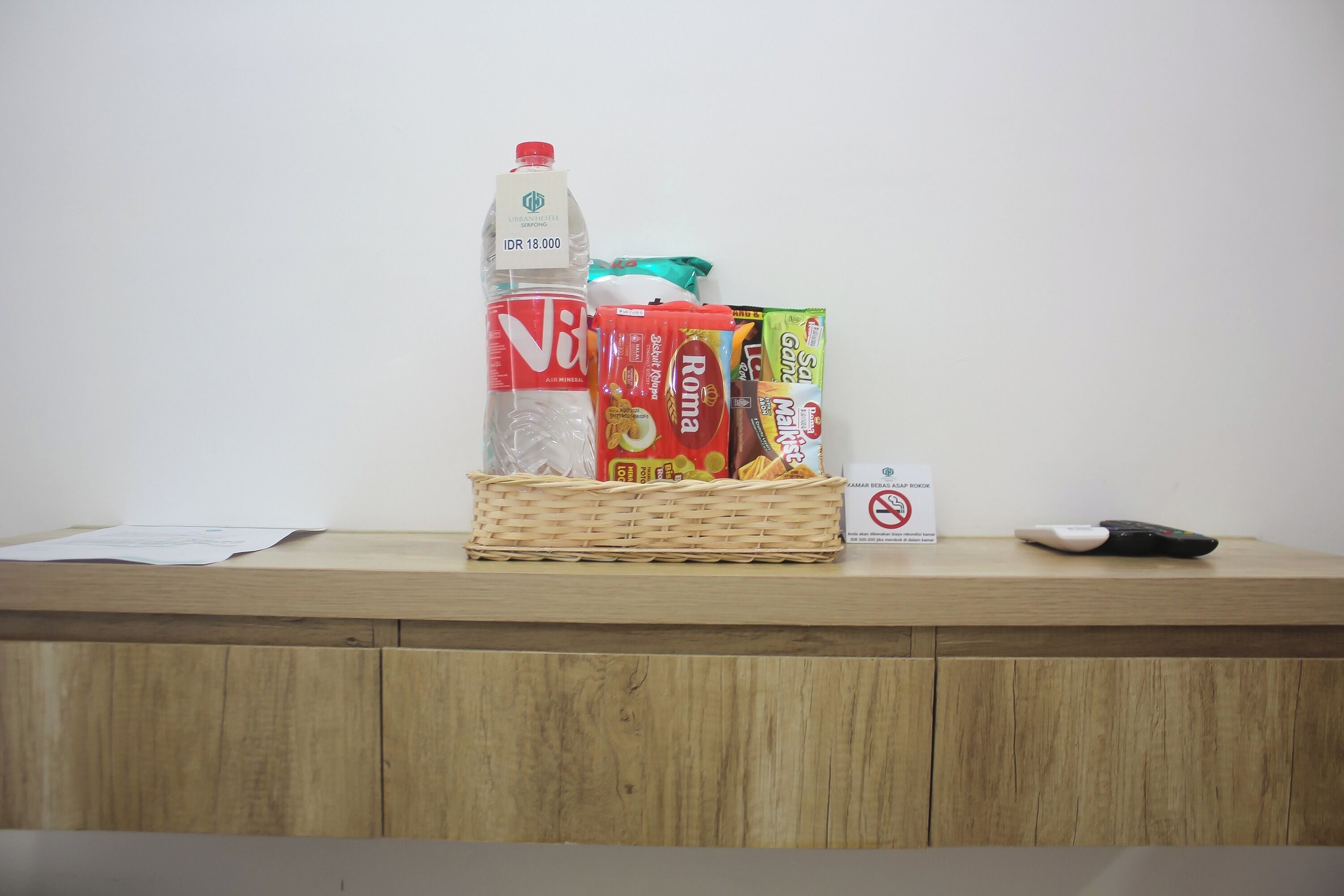 A wooden counter with a basket containing a bottle of water, a box of Nescafé, a box of Roma coffee, a pack of Udon noodles, and a remote control.