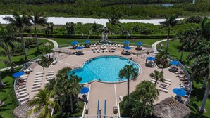 A circular pool with lounge chairs and umbrellas, surrounded by palm trees and a view of a water body. - Stunning Southern Beach Views the Whole Family will Love; Heated Pool & WiFi ~Walk to Dining & Shops (Marco Island)
