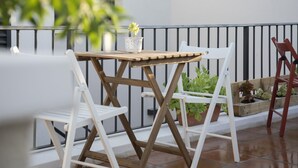 Un balcone con tavolo in legno, sedie bianche, piante in vaso e ringhiera in metallo.
