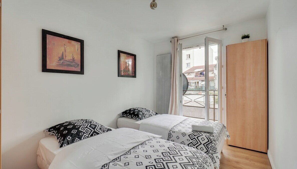 Une chambre avec deux lits, une armoire en bois et un balcon donnant sur des immeubles.