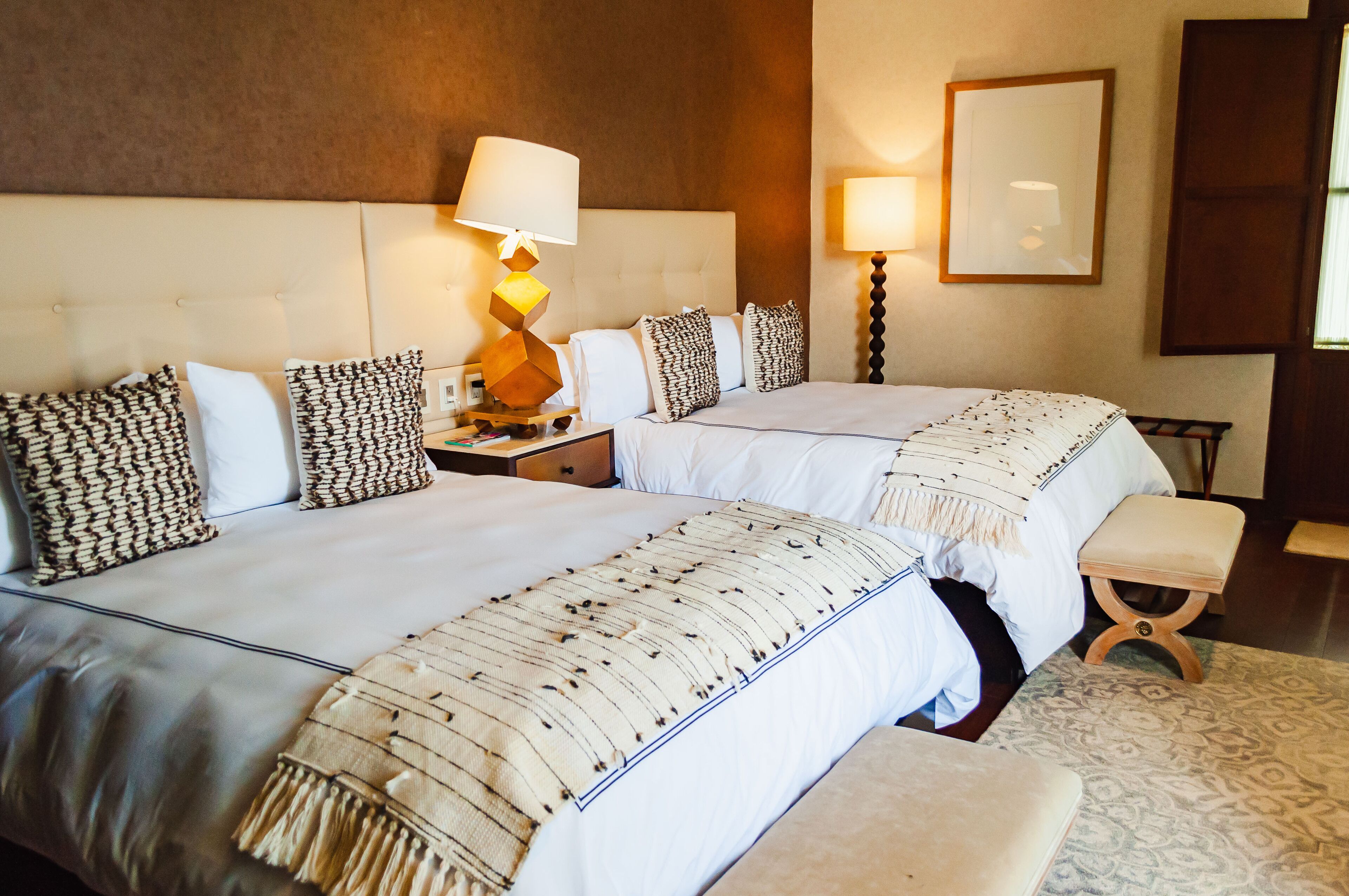 Habitación de hotel con dos camas, una mesita de noche con lámpara, un espejo y una alfombra estampada.