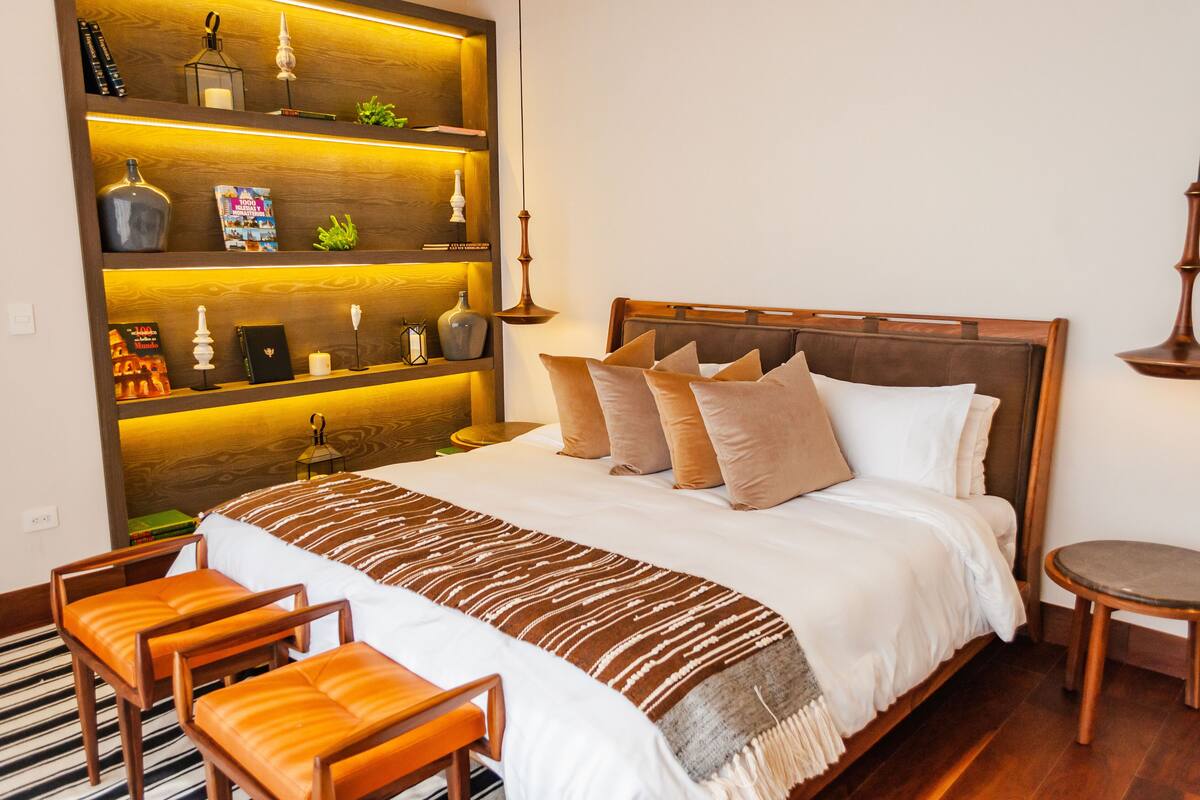 A neatly arranged bedroom with a large bed, a bedside table, a chair, a shelf with decorative items, and a small round table.
