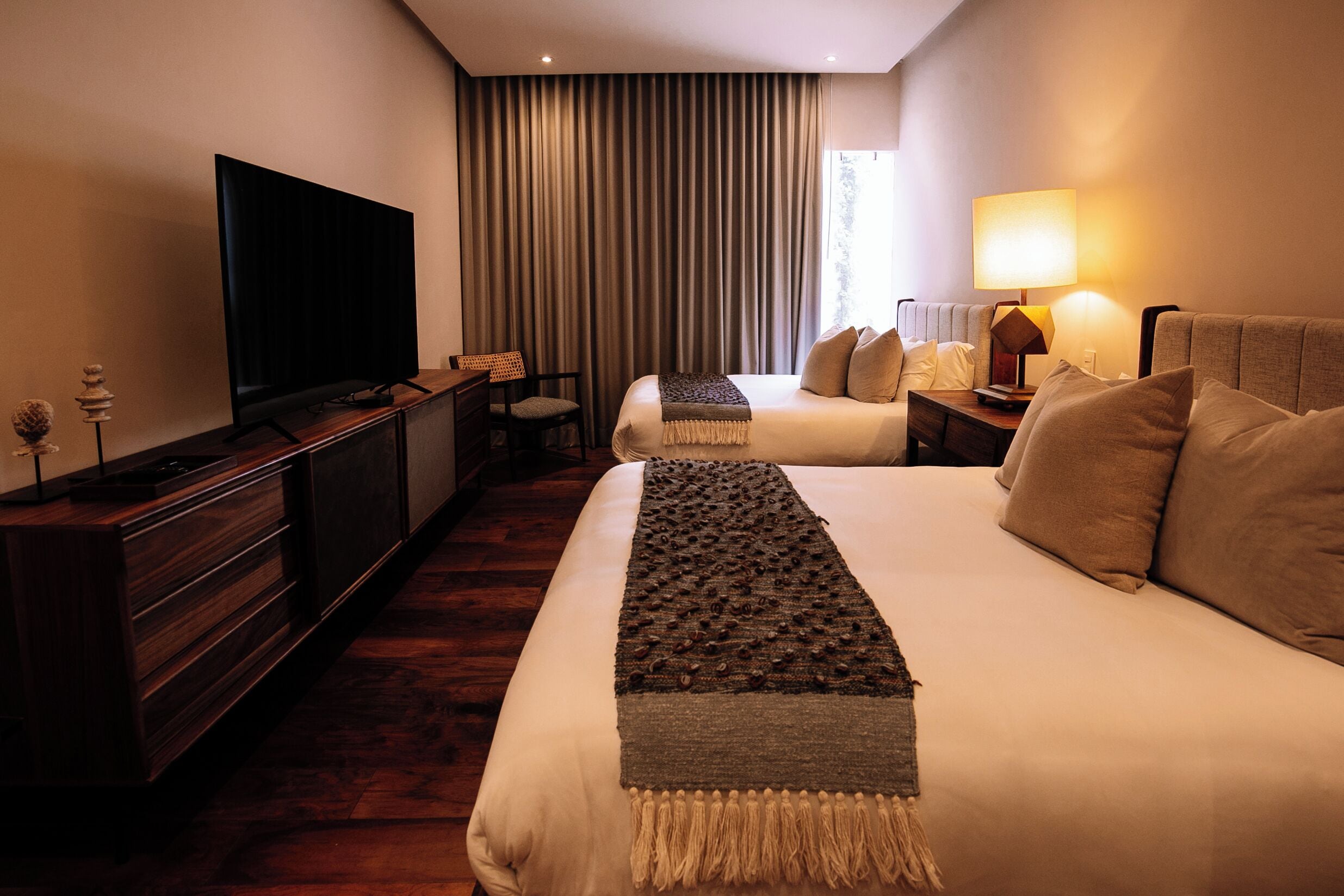 Une chambre d’hôtel dotée d’un grand téléviseur à écran plat, de deux lits avec une literie beige, d’une commode en bois foncé et d’une lampe sur la table de chevet.