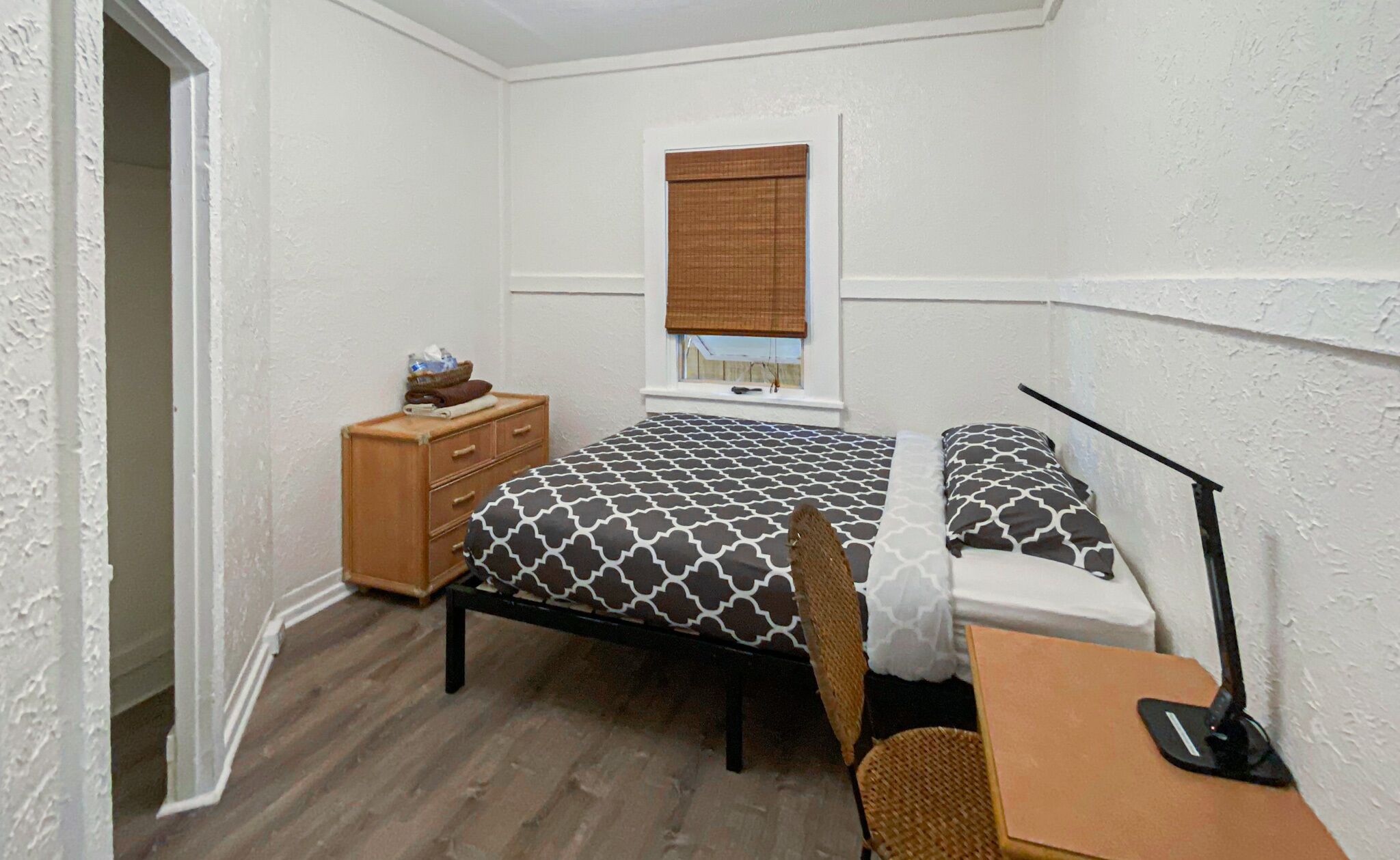 A small, tidy bedroom with a bed, a nightstand, a desk, a lamp, and a window with a blind.