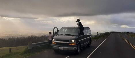 A black van parked on a road with a guardrail on one side and a view of a landscape in the distance.