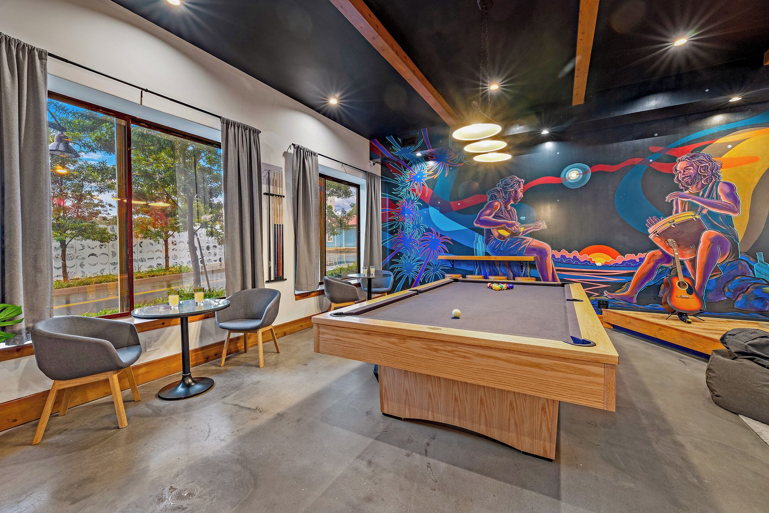 A pool table with a mural, chairs, and a view through large windows.