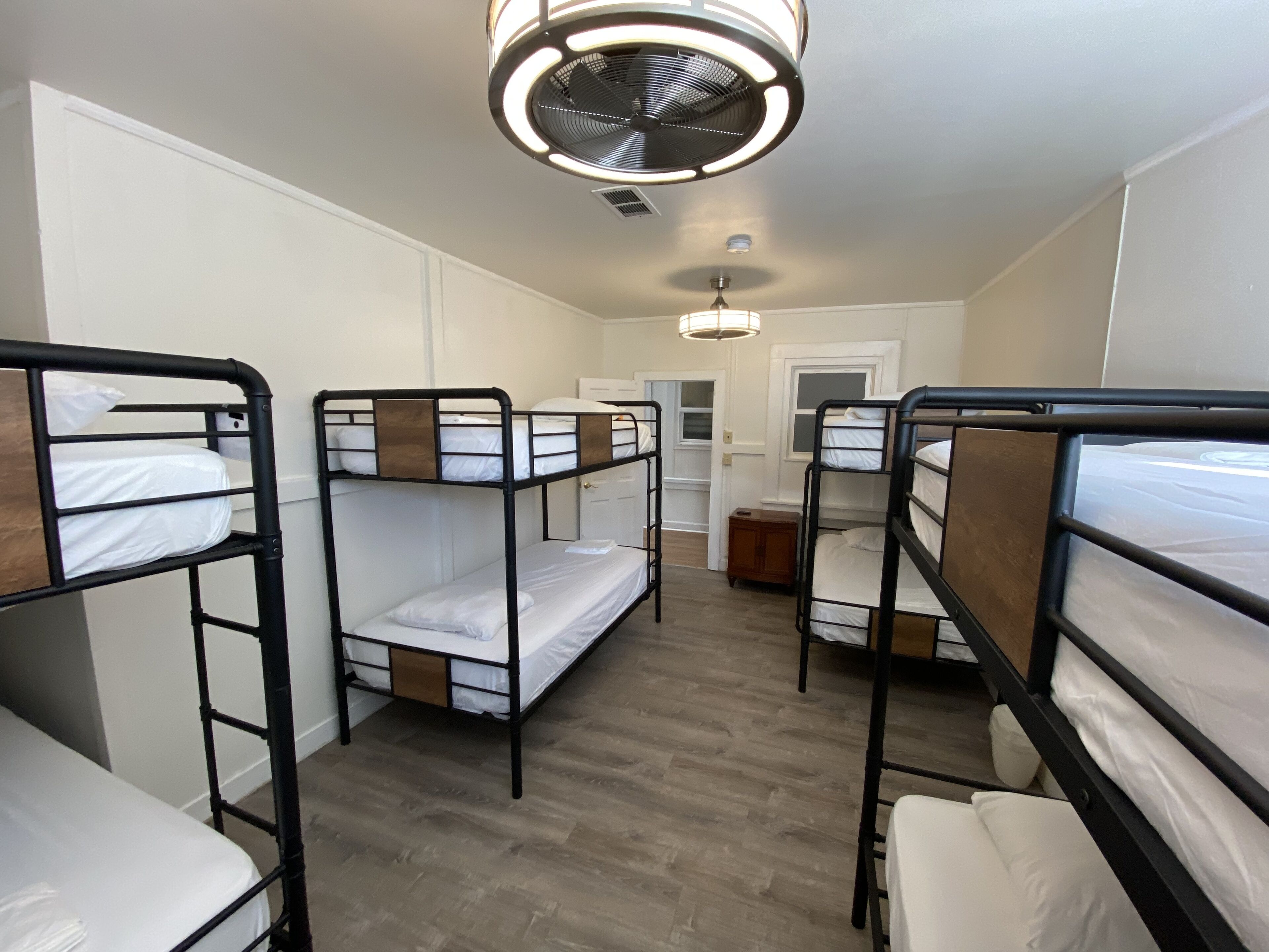 Une chambre de dortoir avec des lits superposés, un ventilateur de plafond et une porte donnant sur une autre pièce.