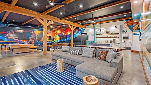 Een moderne woonkamer met een grote grijze hoekbank, houten balken en een kleurrijke muurschildering.