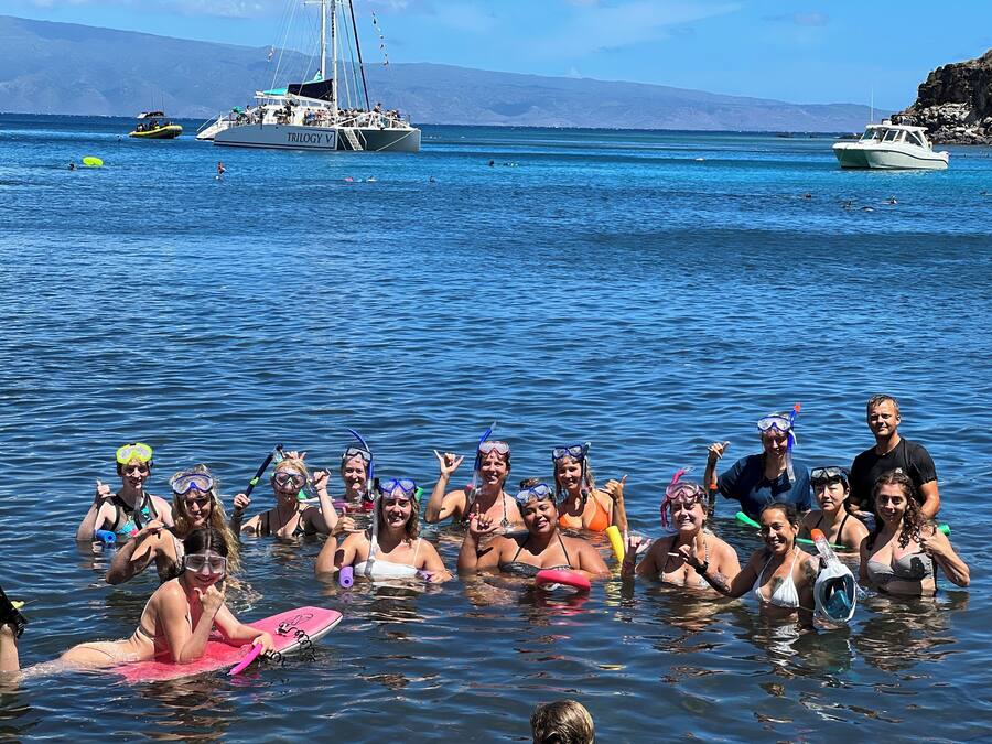 A group of people in a clear blue sea with boats in the background.