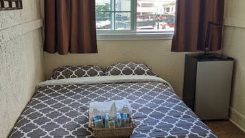 Una habitación de hotel con una cama, una ventana con cortinas, un escritorio pequeño y una cesta de artículos de aseo sobre la cama.