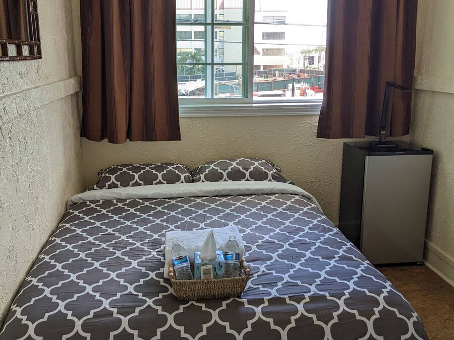 Una habitación de hotel con una cama, una ventana con cortinas, un escritorio pequeño y una cesta de artículos de aseo sobre la cama.