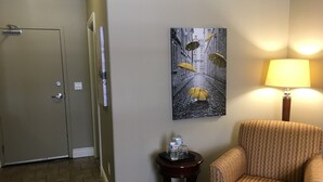 Een hotelkamer met een deur, een klein tafeltje met een fles en glazen, een gestreept fauteuil en een schilderij van paraplu's aan de muur.