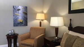 Een hotelkamer met een gestreept fauteuil, een bank, een nachtkastje met een lamp, een tafeltje met flessen en een schilderij van paraplu's aan de muur.