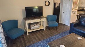 Een woonkamer met een flatscreen televisie, twee blauwe fauteuils, een houten salontafel en een keuken in de achtergrond.