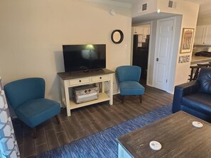 Un salon équipé d’un téléviseur à écran plat, de deux fauteuils bleus et d’une table basse en bois, avec un coin cuisine à l’arrière-plan.